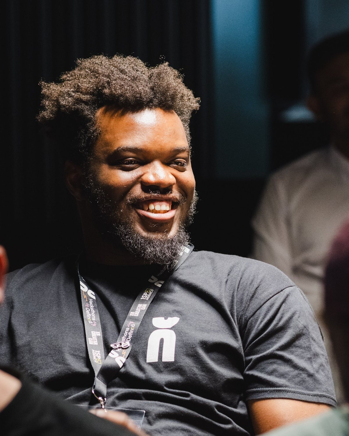 Tolu Aina laughing in a black t-shirt at the XR Creator Challenge, lanyard on, others seated around
